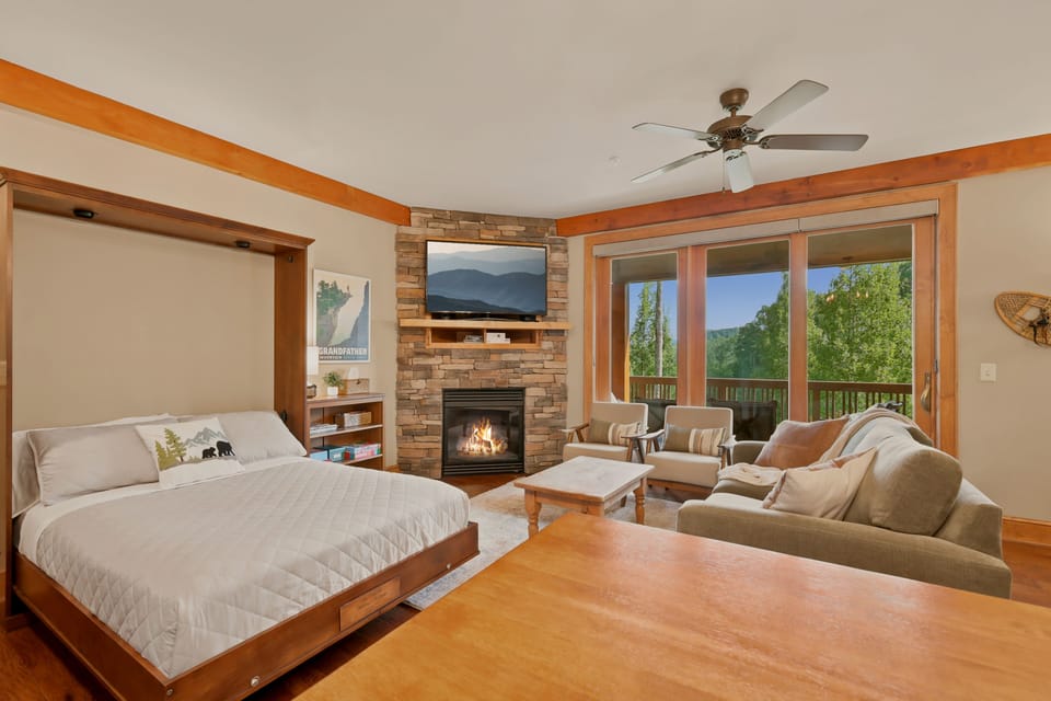 This studio pairs a queen Murphy bed with a dining table and fireside sitting area, floor-to-ceiling windows framing the Blue Ridge treeline and a private balcony just steps away — flexible living for however your group beds down.