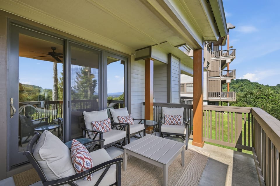 Swivel-glide chairs with patterned pillows circle a slatted coffee table on this covered balcony, complete with its own ceiling fan and a long view over the lawn toward distant ridgelines beyond the neighboring buildings.