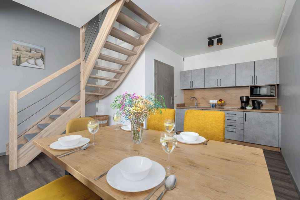 A large wooden dining table is surrounded by vibrant yellow chairs in an open-plan living space. In the background, a wooden staircase leads to an upper level next to a modern kitchenette.