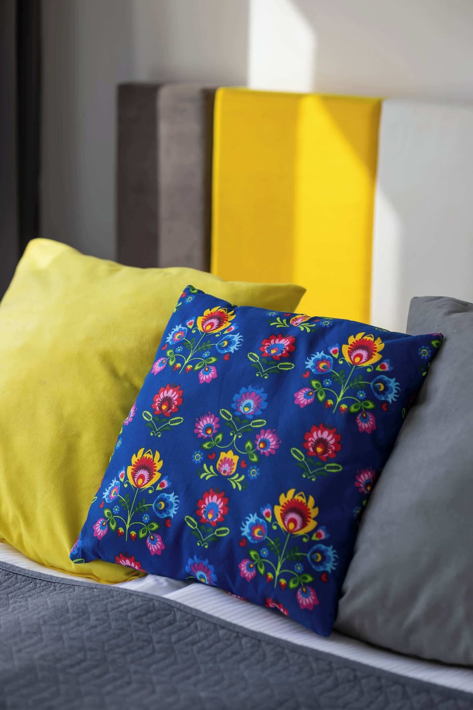 A close-up of a bed with colorful patterned pillows on a gray blanket. The bright yellow and multicolored cushions stand out against the neutral bedding. The detail highlights the decorative style of the bedroom.