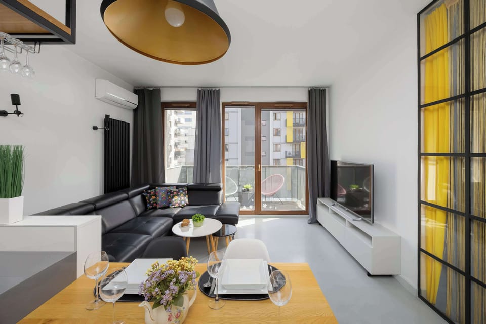 A stylish lounge area featuring a black couch and a coffee table on a patterned rug. Yellow accents appear on doors or wall panels in the background. Decorative lighting hangs from the ceiling above the seating area.
