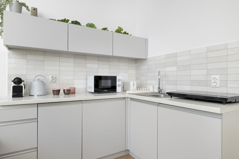 A clean white kitchenette with modern cabinets and open shelves mounted on the wall. Small plants and kitchen items decorate the shelves. The design looks minimalistic and functional.