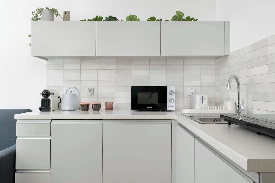 Another angle of the same kitchenette with white cabinets and floating shelves. A microwave and small appliances sit on the countertop. The space appears tidy and contemporary.