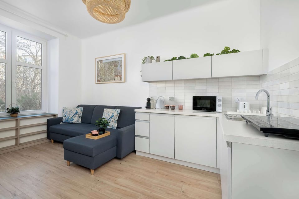 A modern open-plan room with a couch positioned near a small white kitchenett area. The kitchenette cabinets and shelves are minimalistic and clean. Light wooden flooring and white walls create a bright and simple interior.