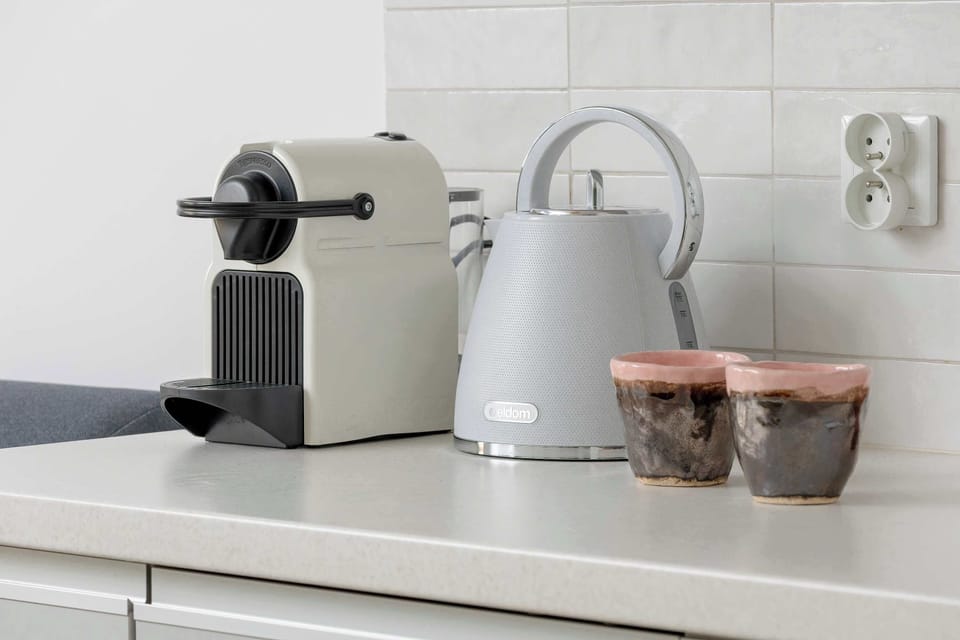 A kitchenette countertop with a coffee machine and cups arranged neatly. The appliances sit on a white counter against a light wall. The setup suggests a convenient coffee corner.