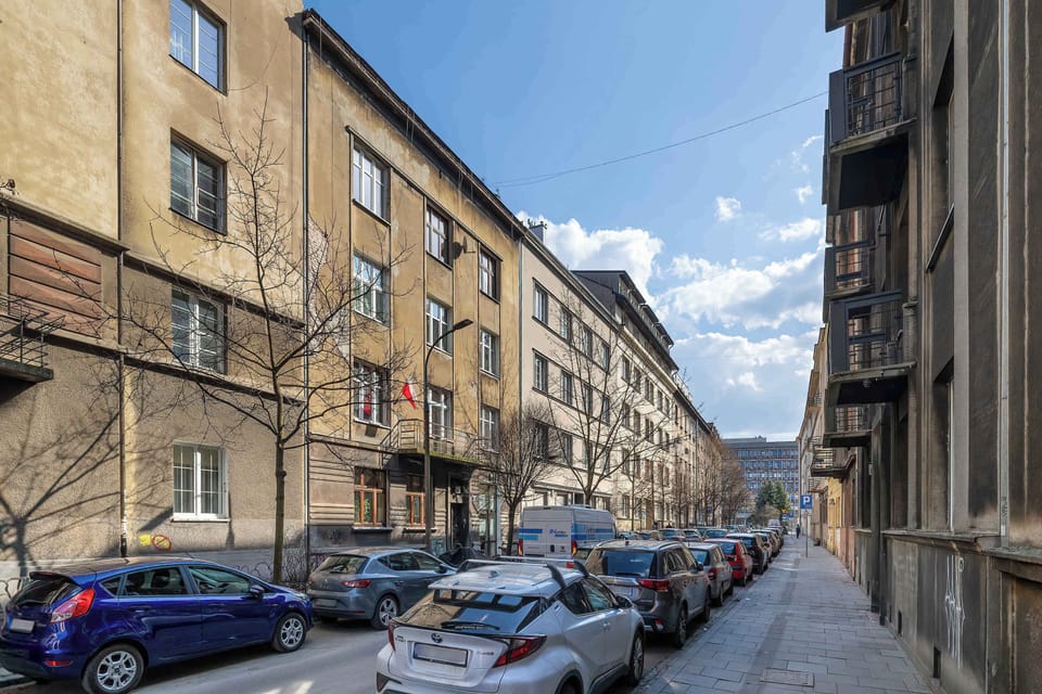 An exterior street view of a residential building with several parked cars along the road. The building has multiple floors and classic European-style architecture. 