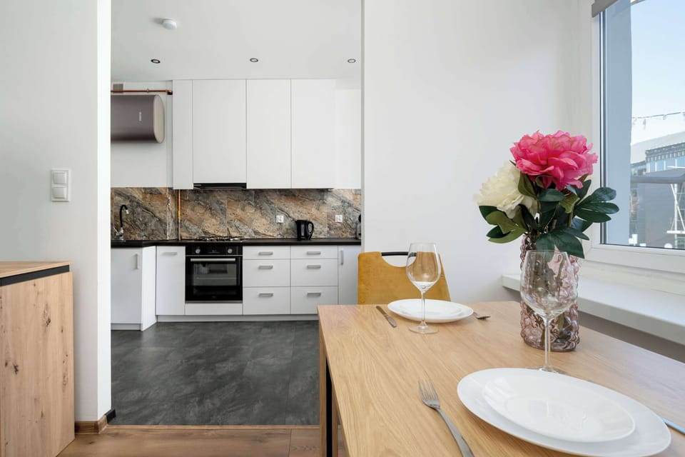 A kitchenette counter with a wooden tabletop extension used as a dining spot. A plate and cutlery are arranged on the table, and a vase with pink flowers adds color to the scene. White cabinets and a marble-style backsplash are visible behind it.