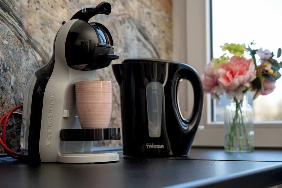 A close-up of a kitchenette counter with an electric kettle and coffee maker. The appliances sit on a dark countertop beside a small vase with flowers. The setup suggests a convenient spot for making coffee or tea.