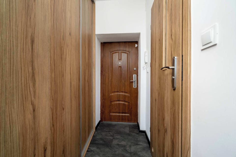 A hallway entrance area with wooden doors and a dark tiled floor. The walls are white, giving the corridor a bright and tidy look. The entrance door is directly visible at the end.