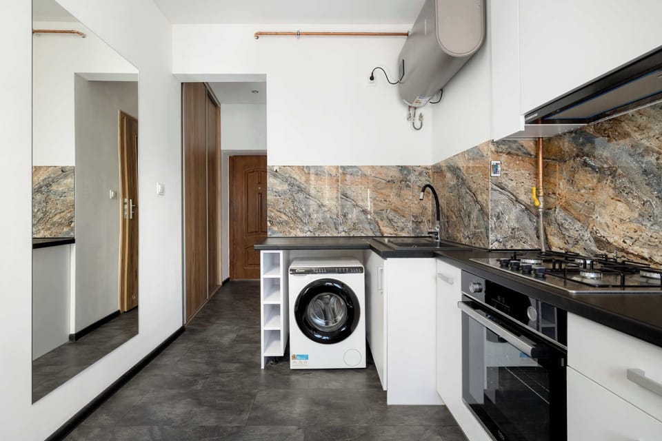 Another angle of the same kitchenette shows more of the appliances and countertop space. The washing machine, stove, and overhead cabinets are clearly visible. The design looks modern with white surfaces and dark countertops.