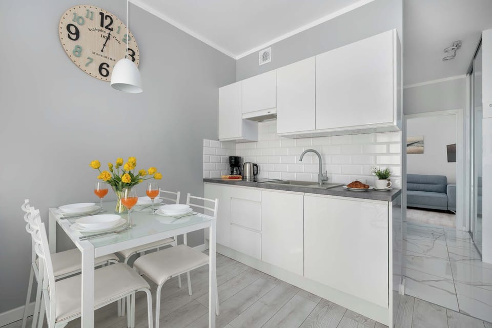 This view focuses on a white dining table with four chairs positioned close to a modern kitchen corner. A clock and a light fixture hang above.