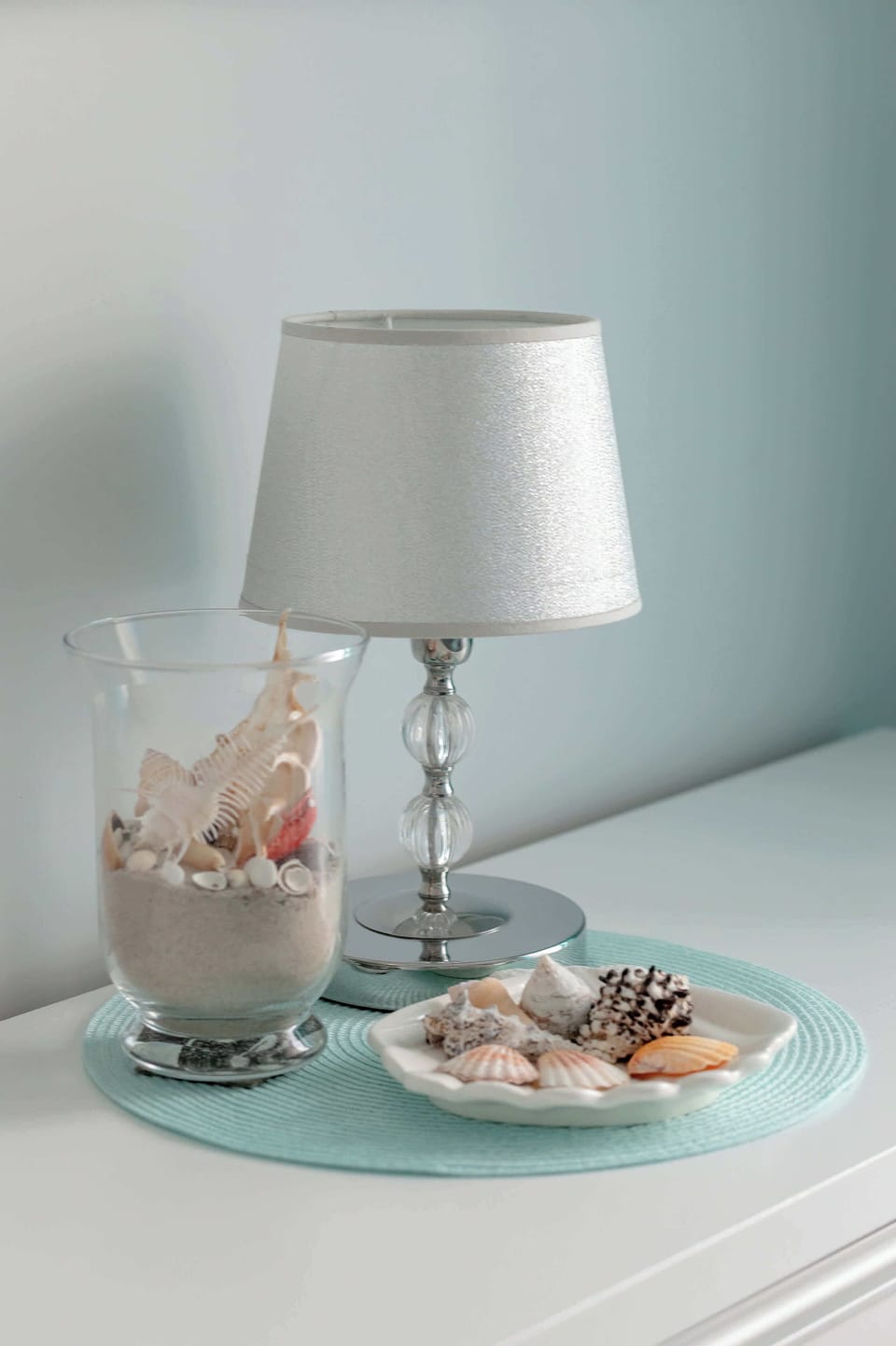 A modern lamp with a beige shade sits on a white console table. Next to it are decorative items, including a plate with various small objects.