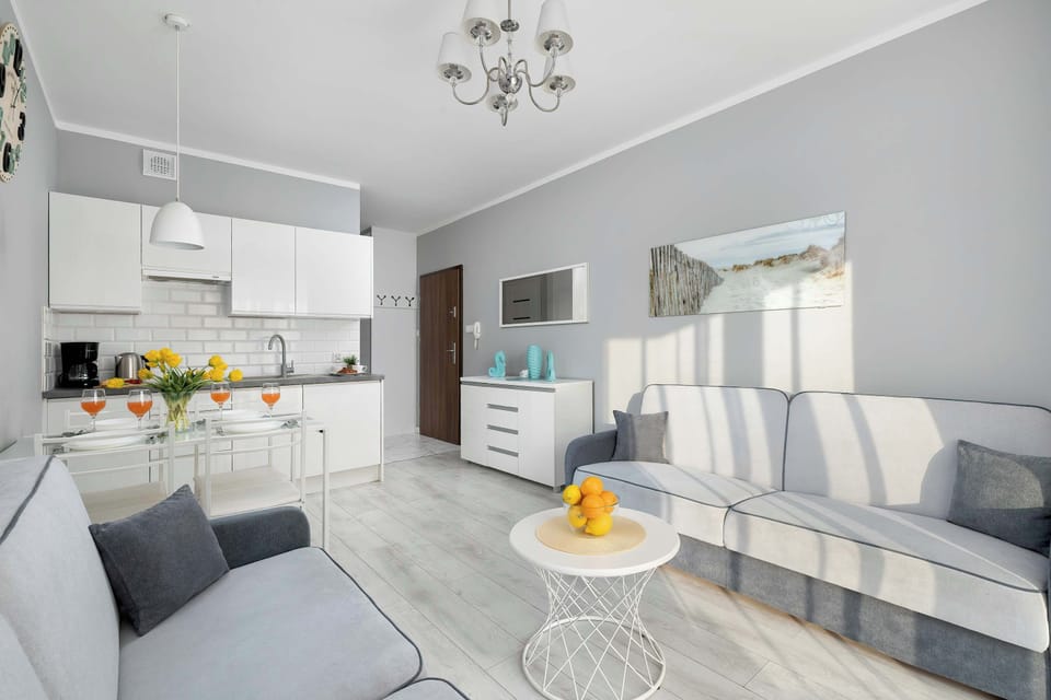 The grey L-shaped sofa and a glass-topped dining table are clearly visible in this open-plan living and kitchen area. The kitchen features white cabinets and a dark countertop.