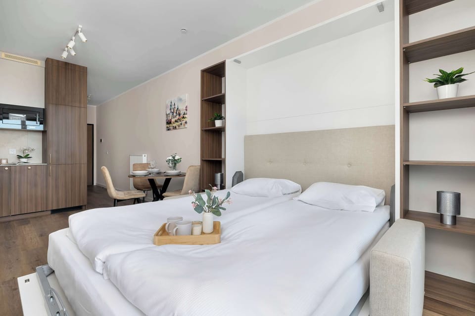 A bedroom corner displays a bed with neutral bedding and built-in shelves along the wall. Small decorative items and soft lighting add a cozy touch.