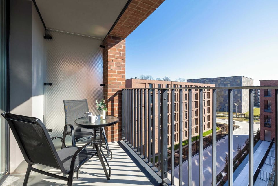 A balcony with two chairs and a small table overlooks neighboring apartment buildings. Metal railings and a clear view make it a pleasant outdoor spot.