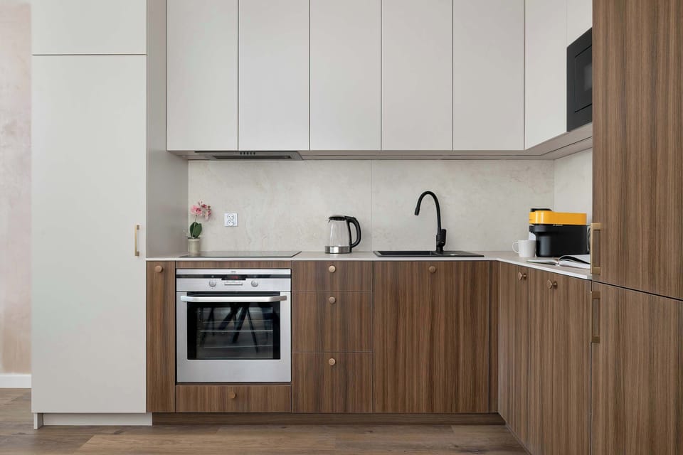 A modern kitchenette with white upper cabinets and wooden lower cabinets stands against a light wall. Built-in appliances and a compact layout make the space practical and efficient.