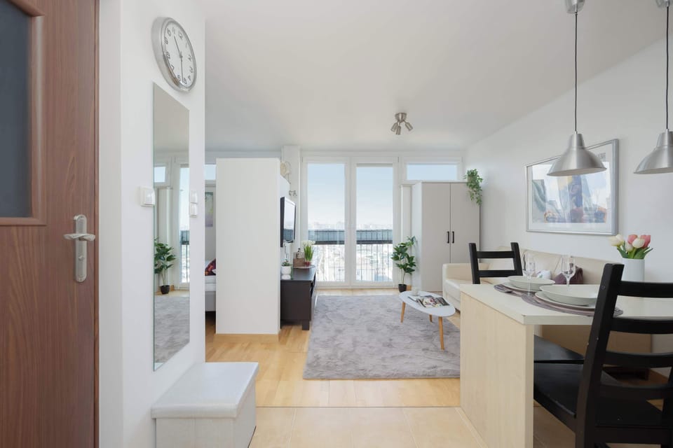 A wide-angle view from the hallway looks past a wall clock and a large mirror toward the sunlit living area. The transition between the functional entry space and the comfortable living zone is seamless and bright.
