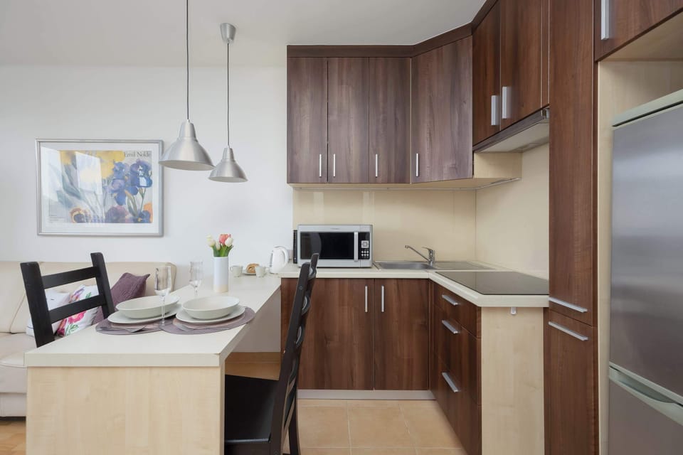 The kitchenette area is designed with dark wood cabinets and integrated appliances, including a silver refrigerator and a microwave. A light-colored dining table with two chairs sits nearby, lit by stylish hanging pendant lamps.