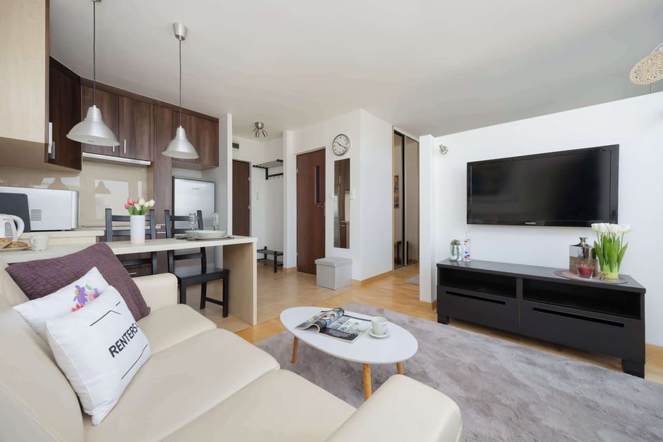 The living area is centered around a large flat-screen TV mounted on a white wall above a black console. A light-colored sofa and an oval coffee table face the screen, offering a perfect spot for entertainment.