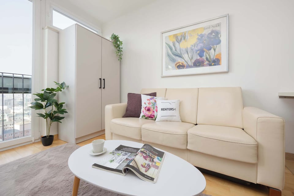 This perspective of the living room highlights a large white wardrobe and a lush green plant standing beside the sofa. An oval coffee table rests on the soft grey rug, creating a central point for relaxation.