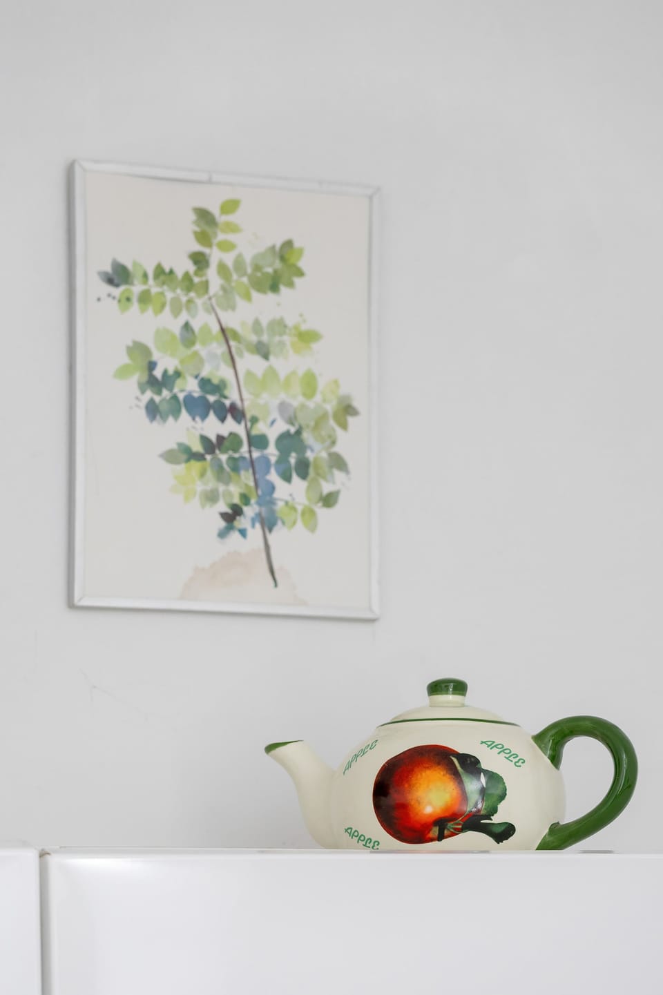 A close-up shot of an artwork featuring green leaves in a white mug is displayed above a decorative green and white ceramic pot. The simple decor elements rest on a white shelf against a plain wall.