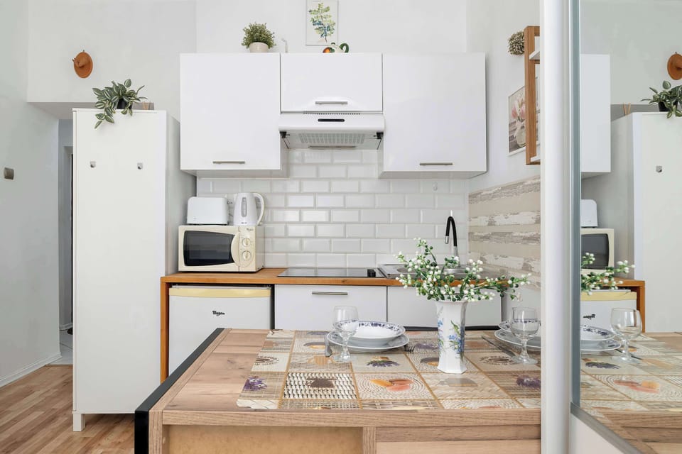 A kitchen-dining area is equipped with sleek white upper cabinets, a light wood counter, and a dining table set for one, complete with a glass vase of faux greenery. The room is modern and clean, showcasing built-in appliances like a microwave.