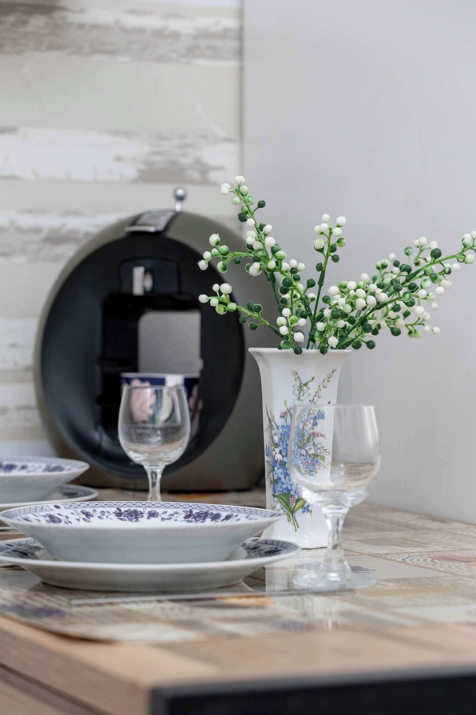 A glass pitcher filled with faux eucalyptus branches sits on a polished dining table, reflecting a modern appliance and kitchen unit in the background. The scene emphasizes minimalist decor in a practical home setting.