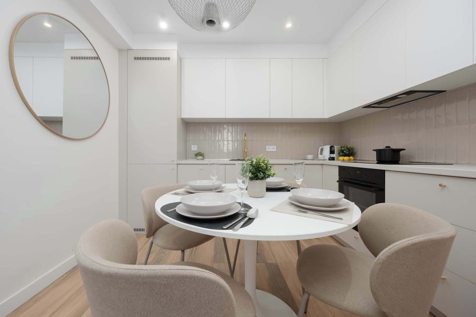 A bright kitchen features a round white table surrounded by elegant beige upholstered chairs. Modern white kitchen cabinetry and minimalist decor create a clean, welcoming atmosphere.