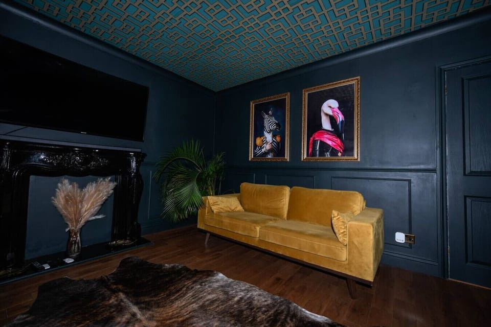 The lounge is arranged with a yellow sofa, a faux-fur rug, framed wall art, and potted plants set against dark-painted walls beside a doorway.