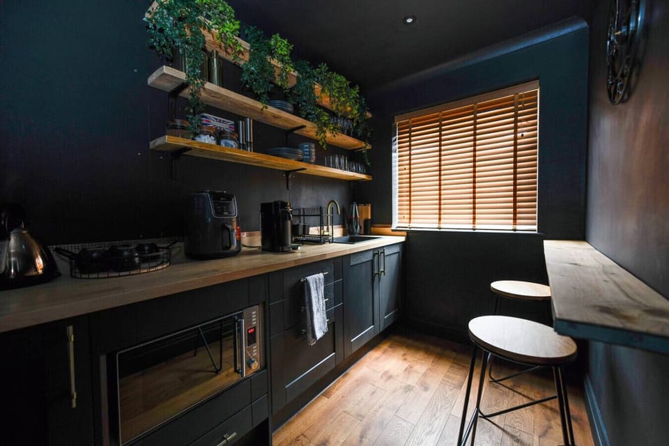 The kitchen area has dark fitted units, open wooden shelving stocked with kitchenware, a bar-height counter with stools, and a wooden-slatted blind at the window.