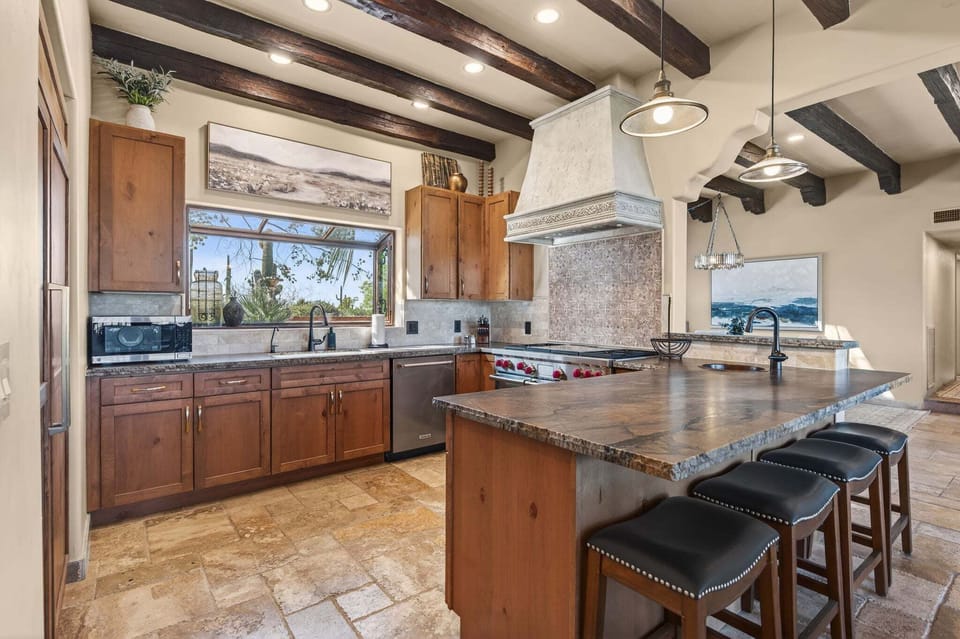 The kitchen includes a broad island, four stools, farmhouse sink, Wolf range, decorative hood, wood cabinetry and garden-facing window.