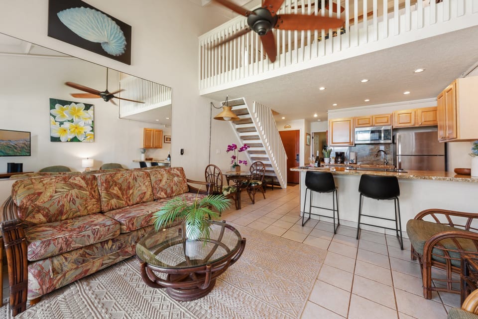 Relax in the spacious, sunlit living area featuring high vaulted ceilings, cozy tropical seating, a modern kitchen, and an open staircase leading up to the loft bedroom
