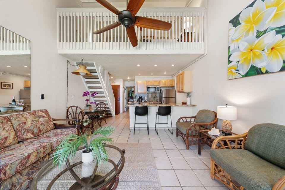 An expansive view of the bright layout, showcasing comfortable tropical seating, high ceilings, kitchen breakfast bar, and open stairs to the loft.