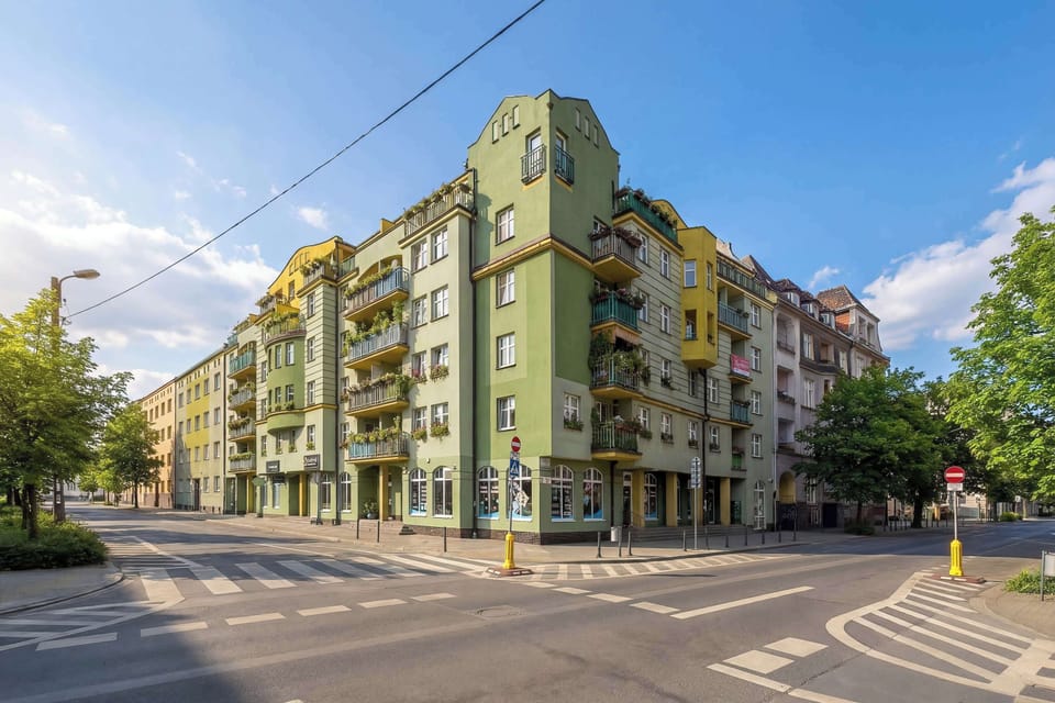 A street corner with a green residential building, balconies, and a clear sky above, showing the surrounding neighborhood.