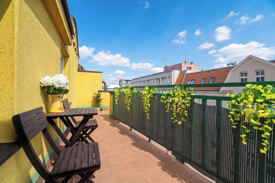 A balcony with yellow walls, outdoor chairs, and a railing, offering a view of nearby buildings.