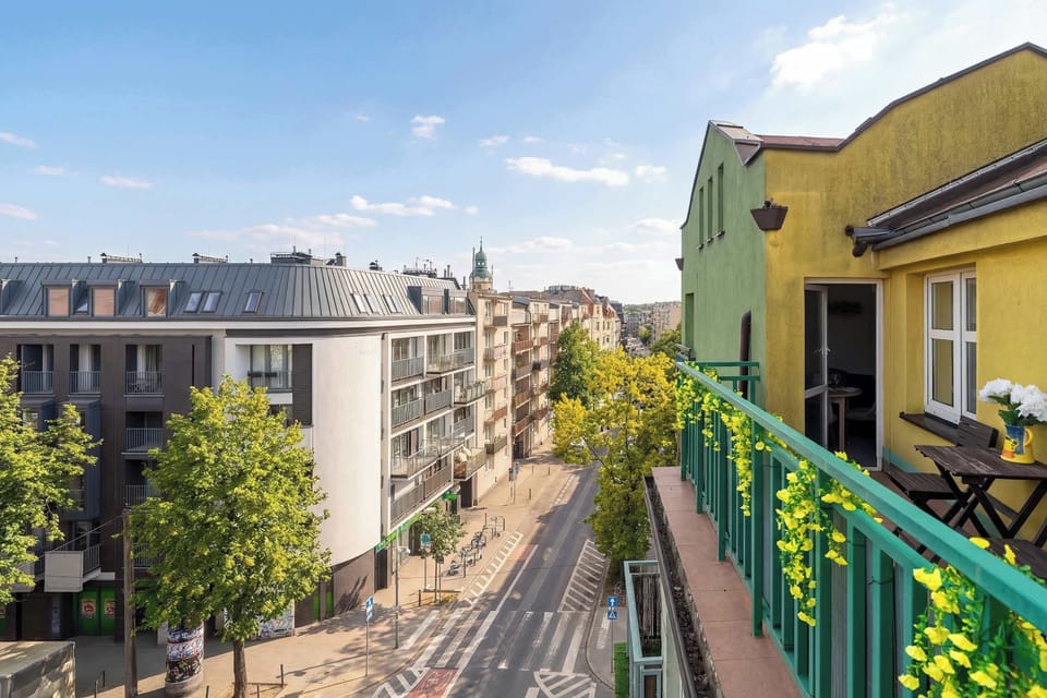 A balcony view overlooking a colorful street with yellow and green buildings, lined with plants and railings.