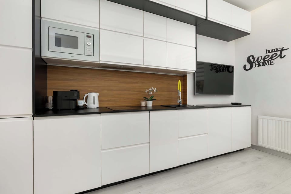 A functional kitchenette with white, handle-less cabinet fronts and wood-effect wall paneling. The built-in units feature a microwave and a minimalist black countertop.