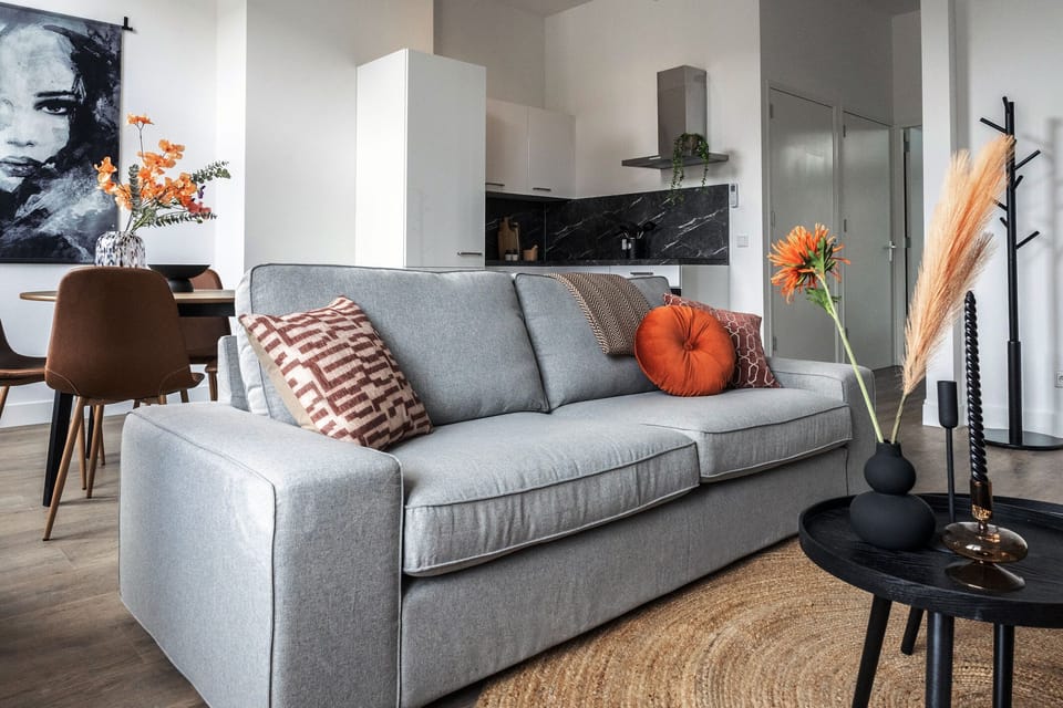 Bright living room with a grey sofa, round black tables, tall windows, cozy lighting, and warm-toned cushions for a welcoming vibe.