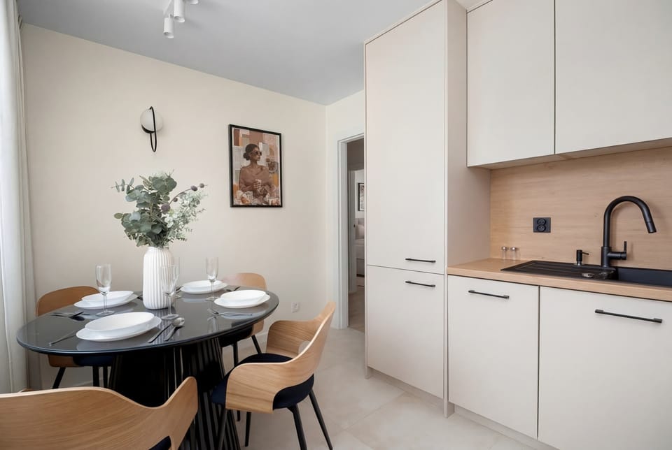 A sleek kitchen with white cabinets and wooden accents. A dining table sits adjacent to the cooking area. The design is simple yet elegant.