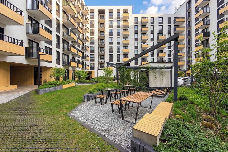 An outdoor recreational area includes wooden benches and tables set amidst landscaped greenery. It provides a peaceful spot for social gatherings within the residential complex.