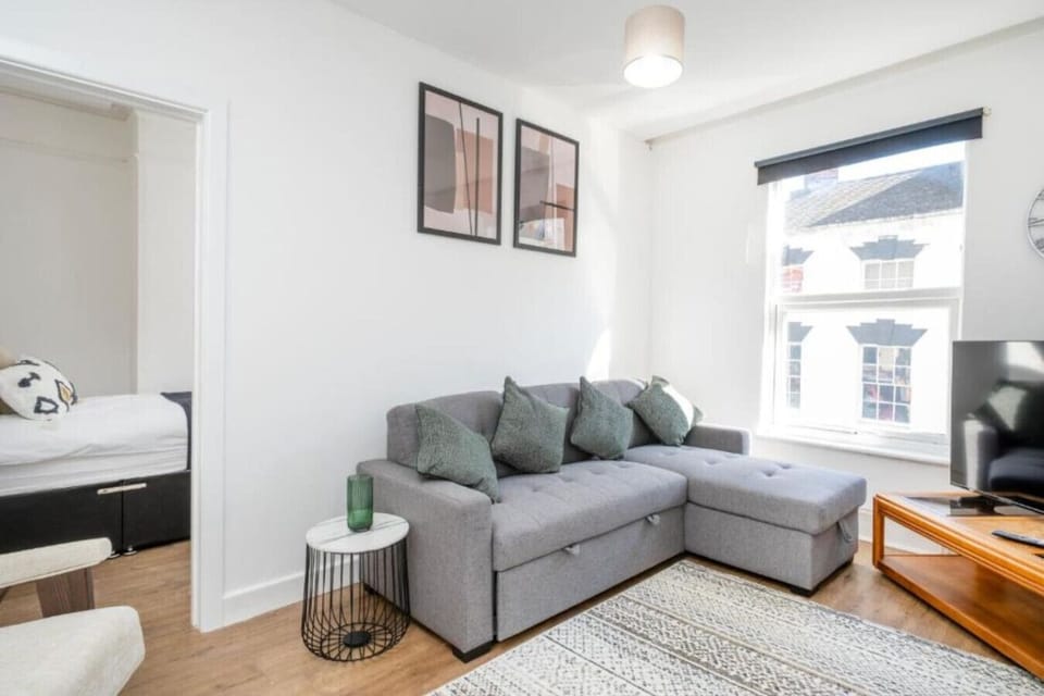 Living area with a grey corner sofa, two framed prints, a round side table, and a sash window.