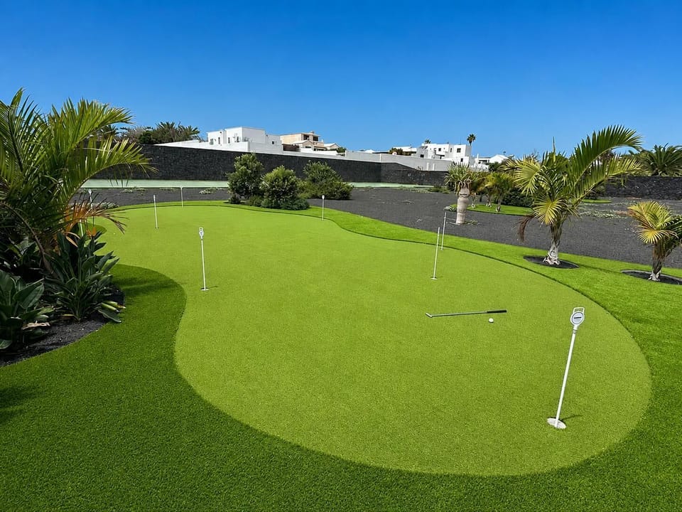 A beautifully manicured private putting green is surrounded by tropical palm trees and a dark volcanic landscape under a bright, partly cloudy sky.