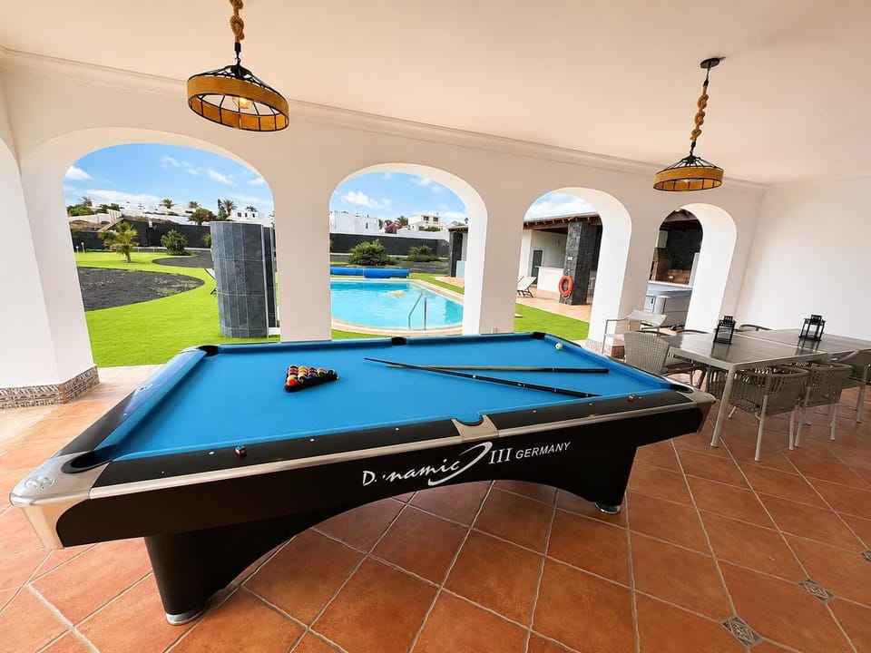 An open-air patio features a professional blue pool table, an outdoor dining set on terracotta tiles, and elegant arches looking out toward a private swimming pool and a lush green lawn.