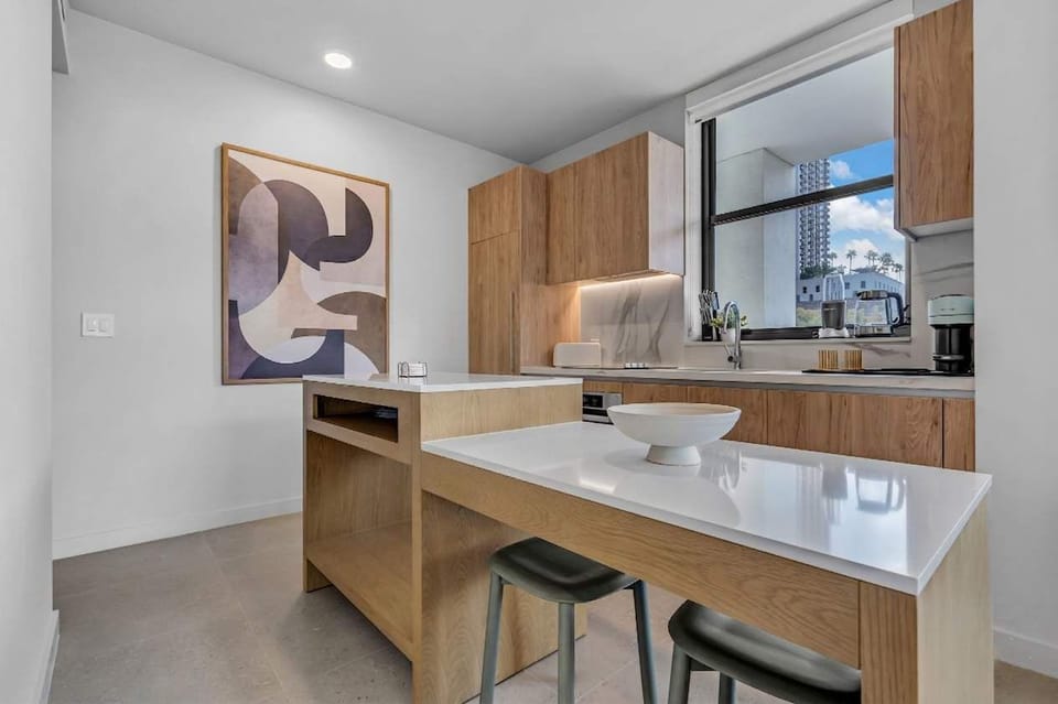 Minimalist chic meets functional design! This bright kitchen space features sleek wood cabinetry, clean white countertops, and a cozy breakfast bar perfect for your morning coffee. Modern art and city views complete this stylish urban oasis.