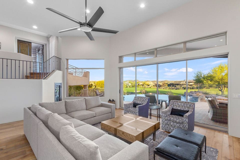 The living room feels bright and open with tall ceilings, a large sectional sofa, and huge windows that frame views of the desert and golf course outside. The space flows naturally toward the outdoor patio.