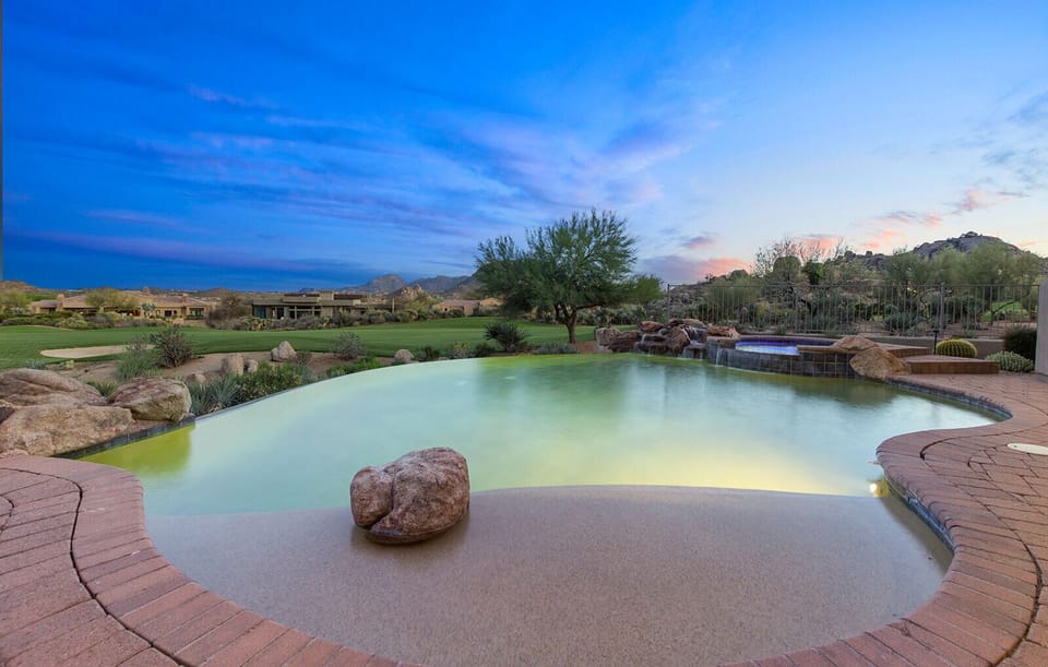 The backyard pool sits right at the edge of the desert landscape, with a smooth beach-style entry and a large stone feature in the water. Beyond the pool, you can see open desert, low hills, and a colorful Arizona sunset sky.