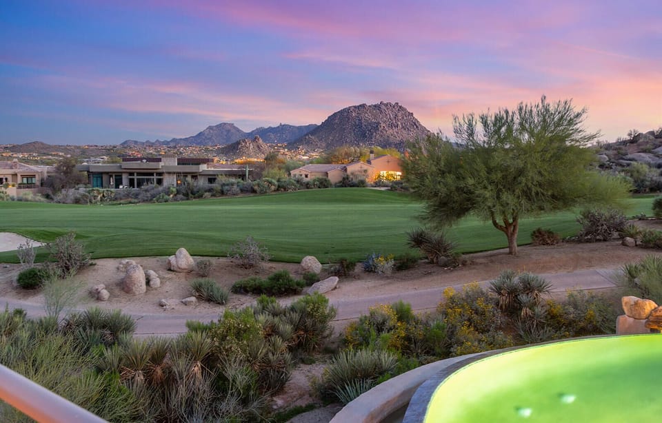 Evening light settles over the backyard as the mountains glow in the distance. Desert plants, natural rock landscaping, and the soft green lawn create a peaceful outdoor setting just as the sun begins to dip.