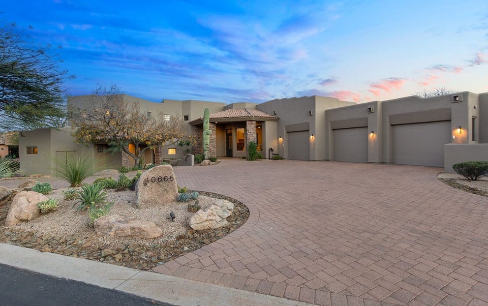 Front exterior of the home with a wide paver driveway, desert landscaping, and a welcoming entryway.