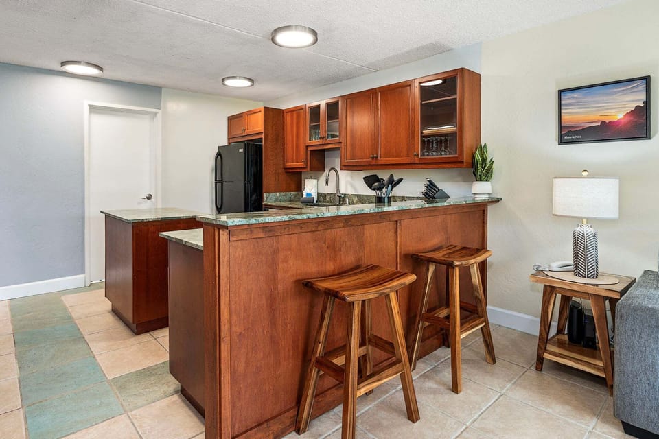 The well-equipped kitchen includes granite countertops, a kitchen island with two bar stools, a cooktop, dishwasher, toaster oven, and an updated coffee bar—perfect for preparing breakfast or enjoying Kona coffee before heading out for the day.