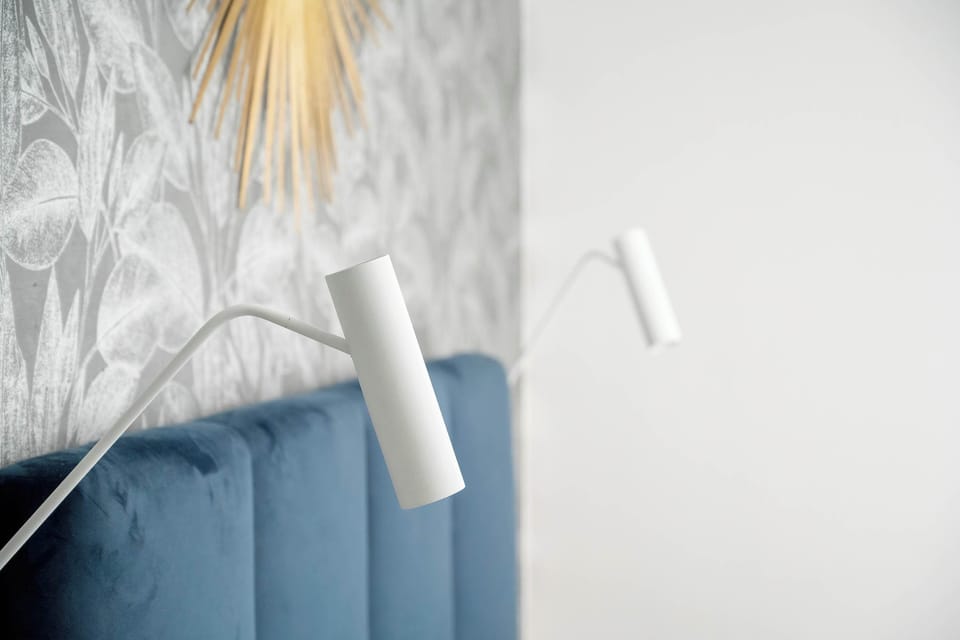 A bedside lighting detail shows a modern white adjustable lamp mounted above the bed. The botanical wallpaper pattern provides a subtle, artistic backdrop for the sleeping area.